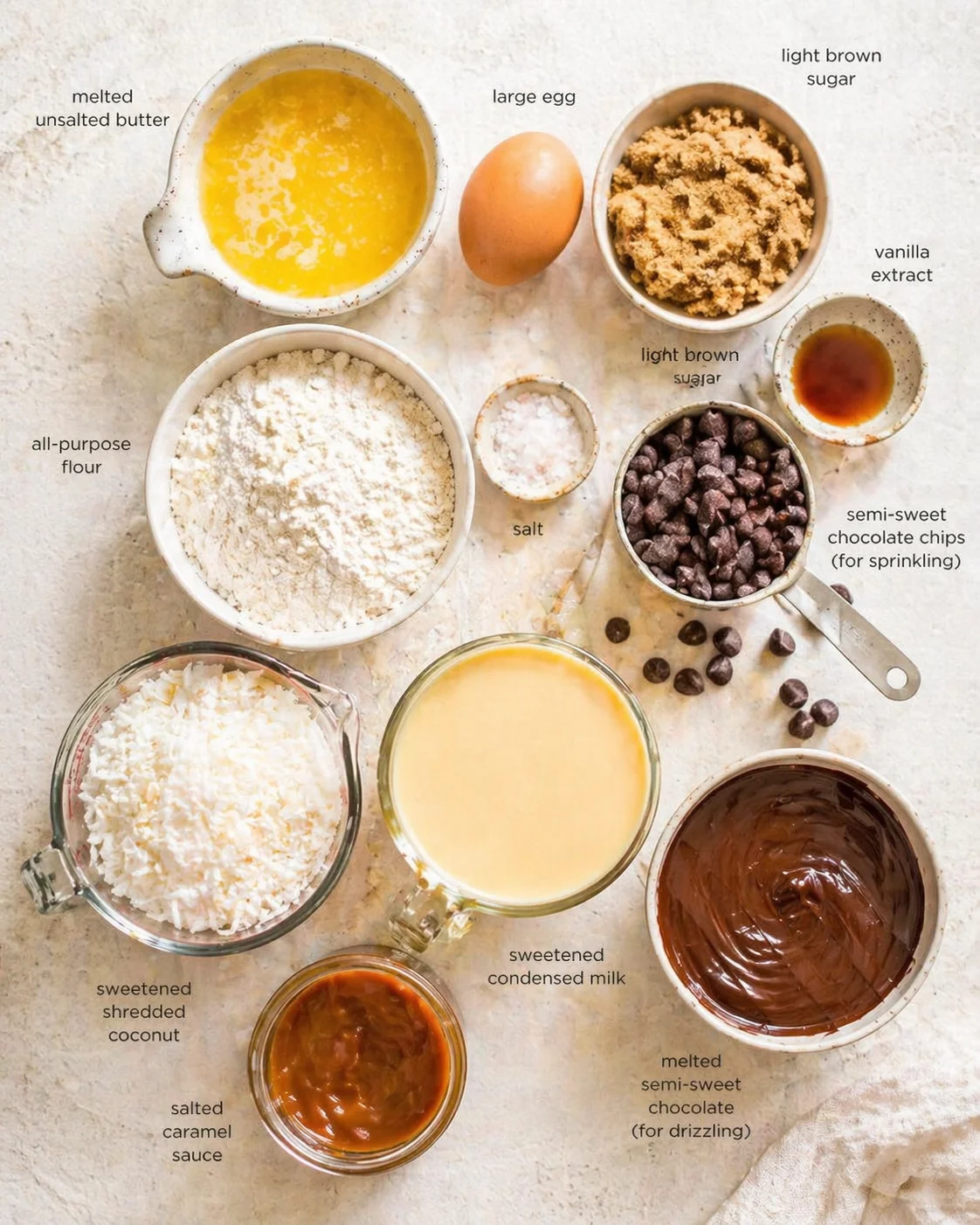 overhead view of butter, egg, sugar, flour, coconut, chocolate, milk and chips arranged on a light textured surface