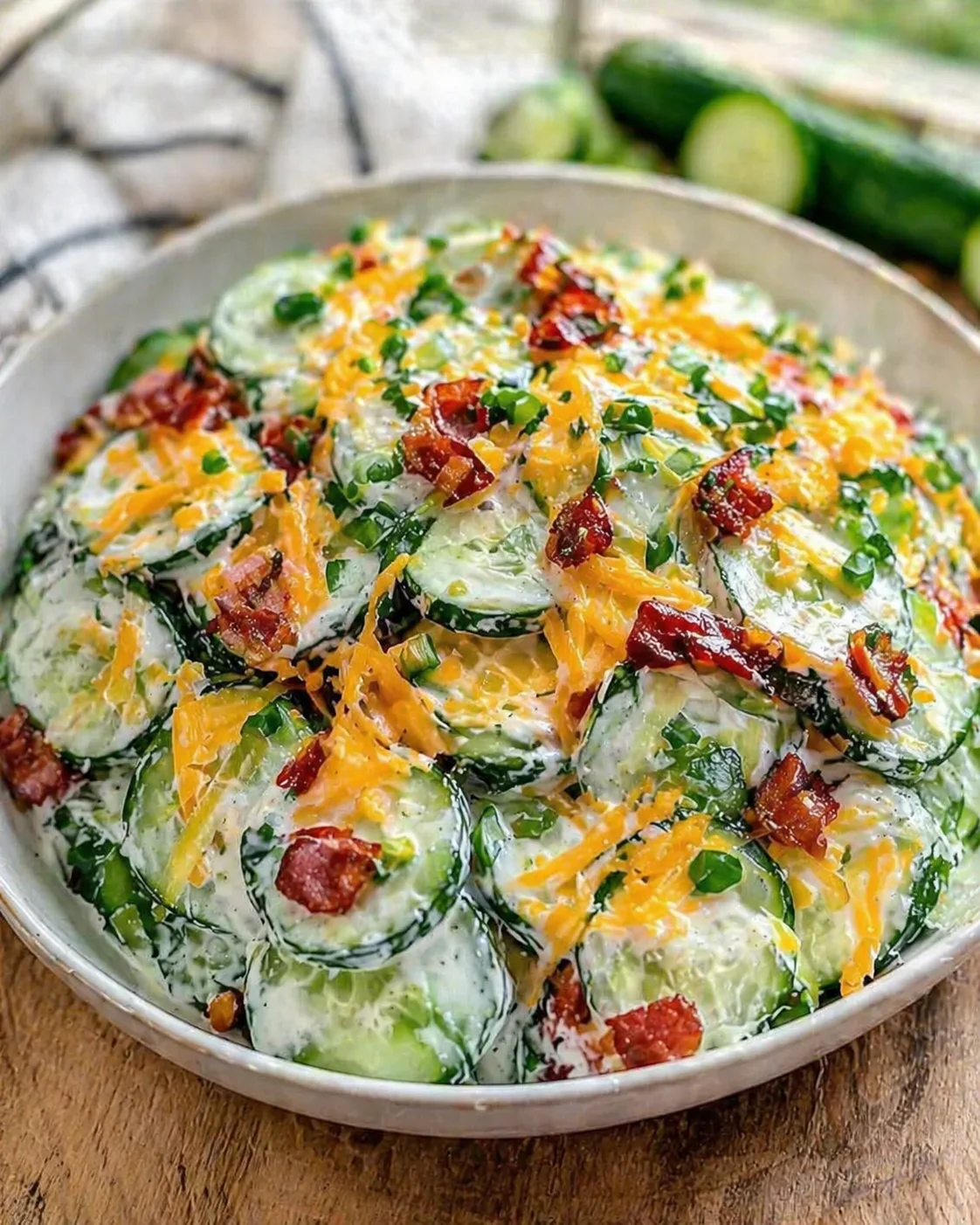 Creamy cucumber salad in a bowl with shredded cheddar, bacon bits, and chopped green onions on a rustic wooden table.