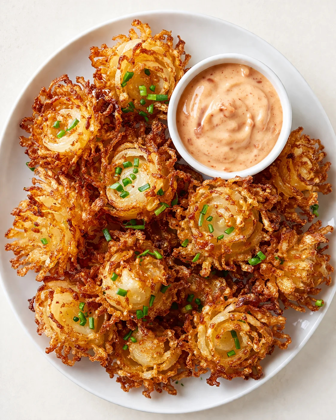 Golden-brown crispy onion rosettes on a white plate with a creamy orange dipping sauce and green chives.
