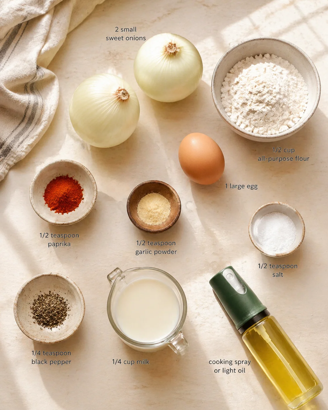 overhead view of onions, egg, flour, milk, spices, and oil spray arranged on a tan kitchen counter