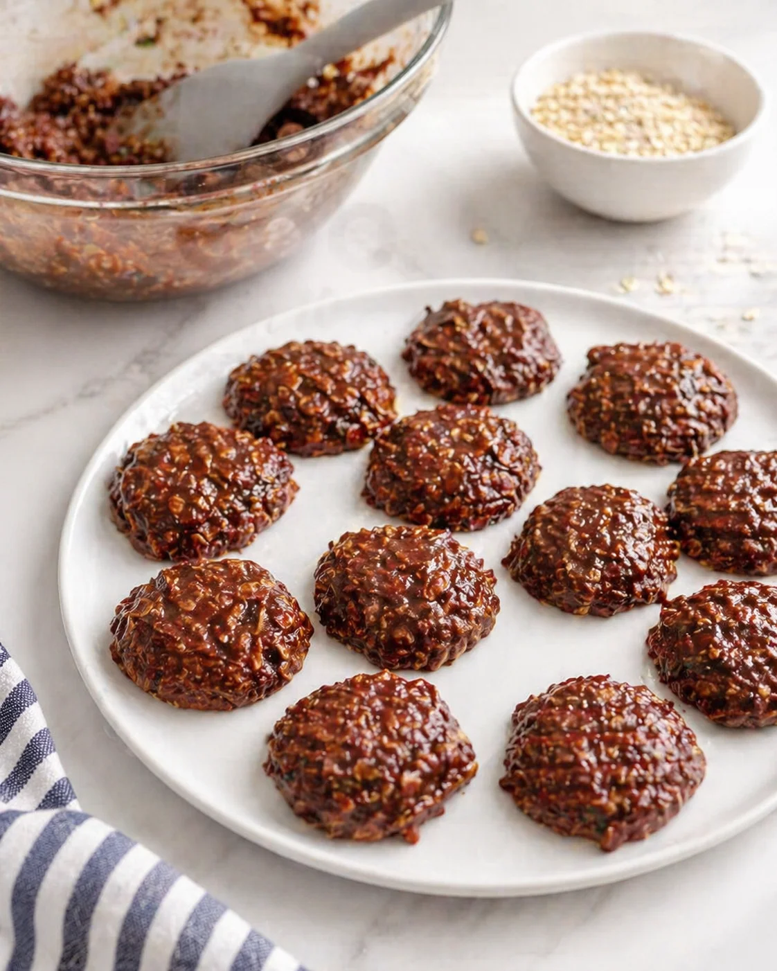 Round no-bake chocolate cookies with chopped nuts on a white plate, glass bowl and sesame seeds nearby.