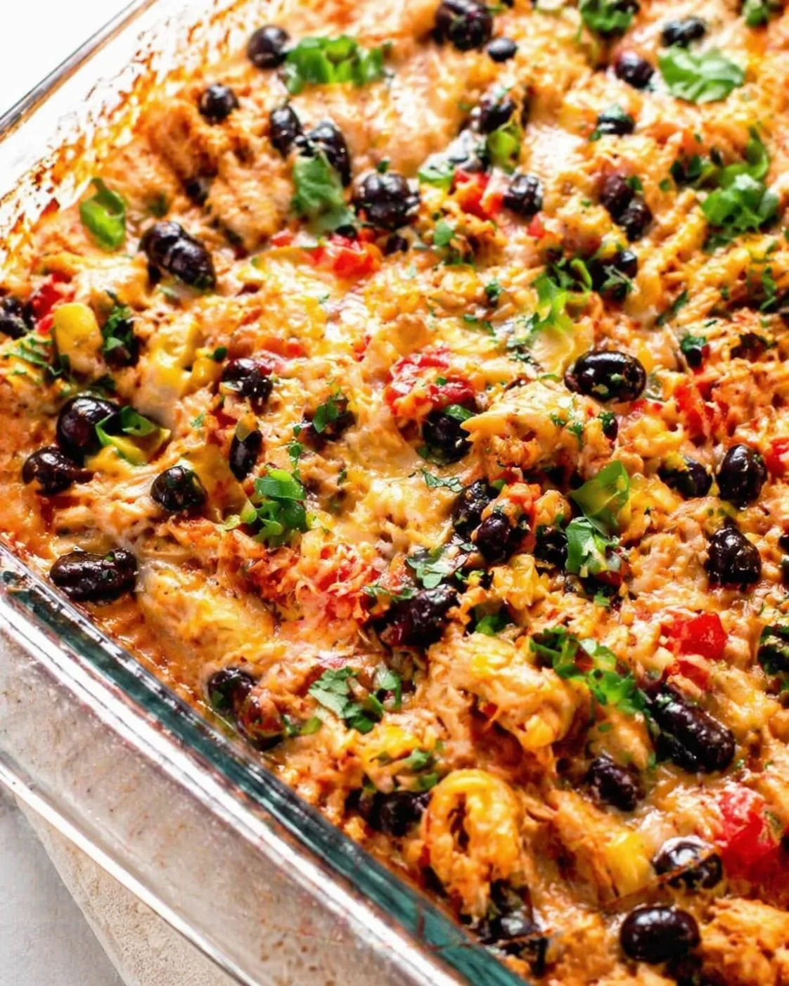 Close-up of a bubbly cheesy casserole in a glass dish, topped with black olives, herbs, and colorful peppers.