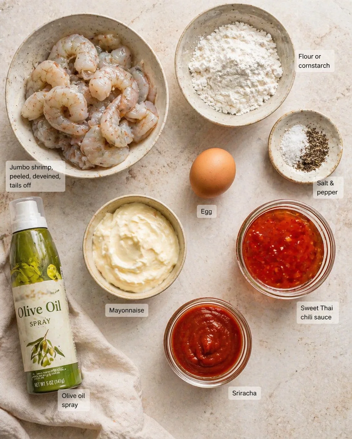 Top-down view of peeled, deveined shrimp with bowls of flour, mayonnaise, sauces, an egg, and olive oil spray.