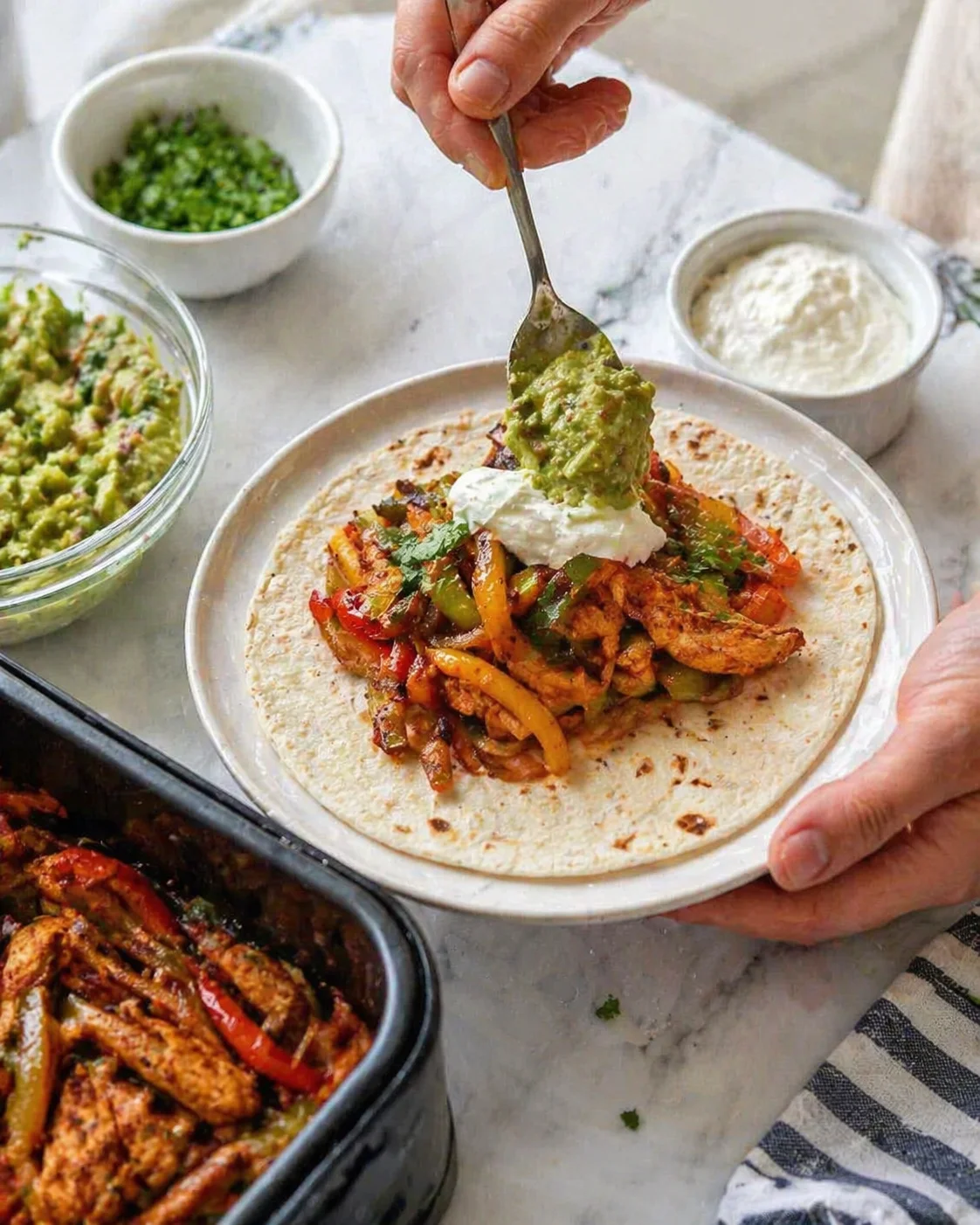 Hand adds spoonful of guacamole to a loaded chicken fajita taco on a white plate.