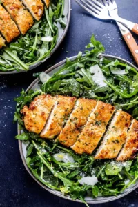 Air fryer breaded chicken breast sliced into strips on a bed of arugula and shaved Parmesan on a plate with another plate of chicken behind it.