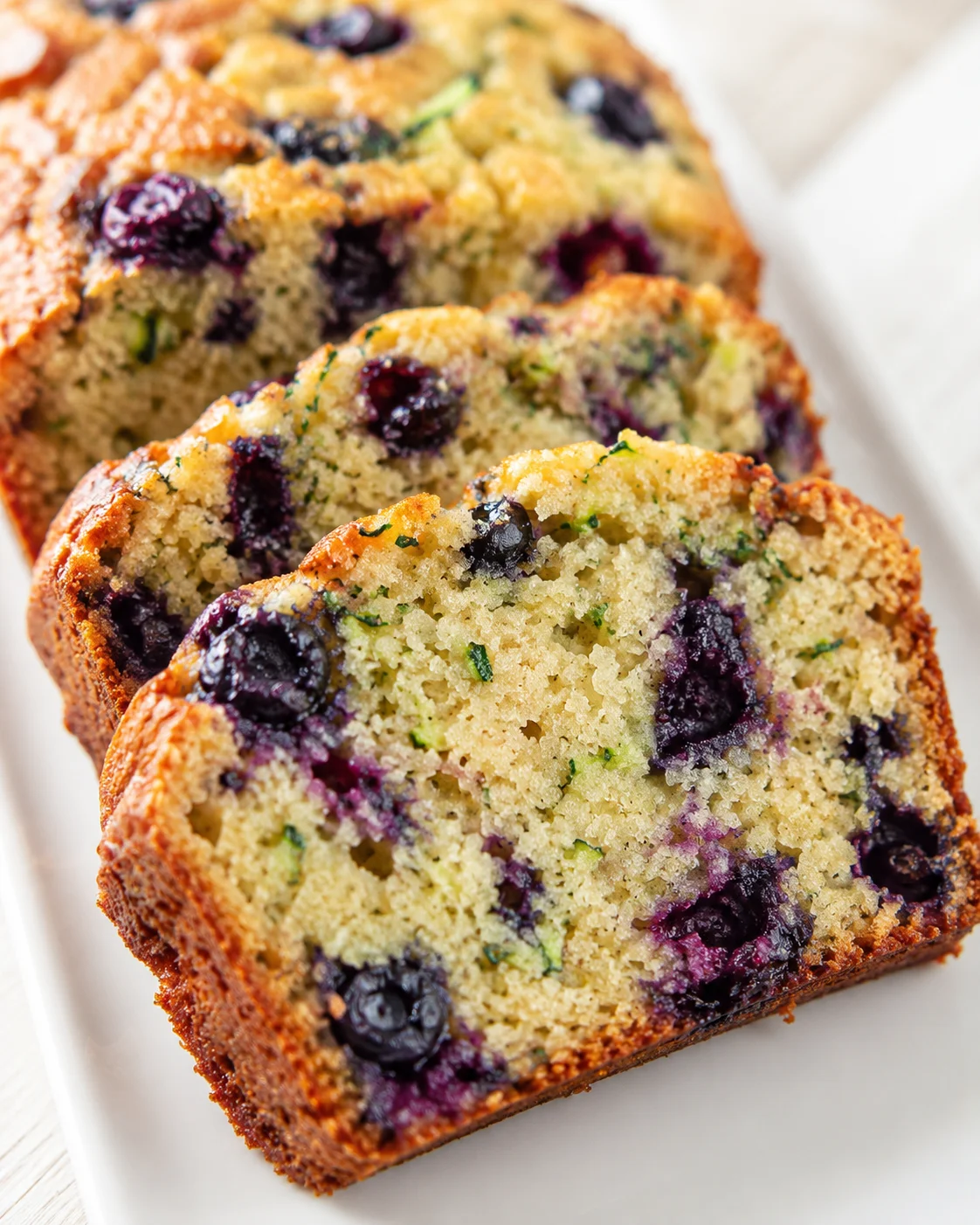 Sliced blueberry loaf cake on a white plate, showing a moist crumb with blueberries and green zest.