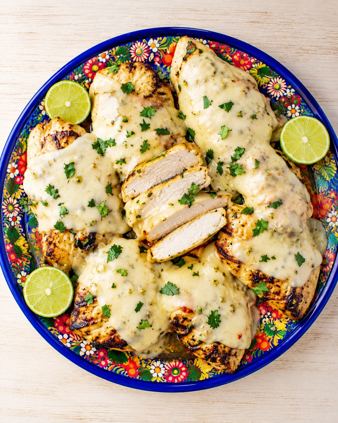 sliced grilled chicken breasts smothered in creamy cheese sauce, garnished with herbs on a bright floral plate with lime wedges