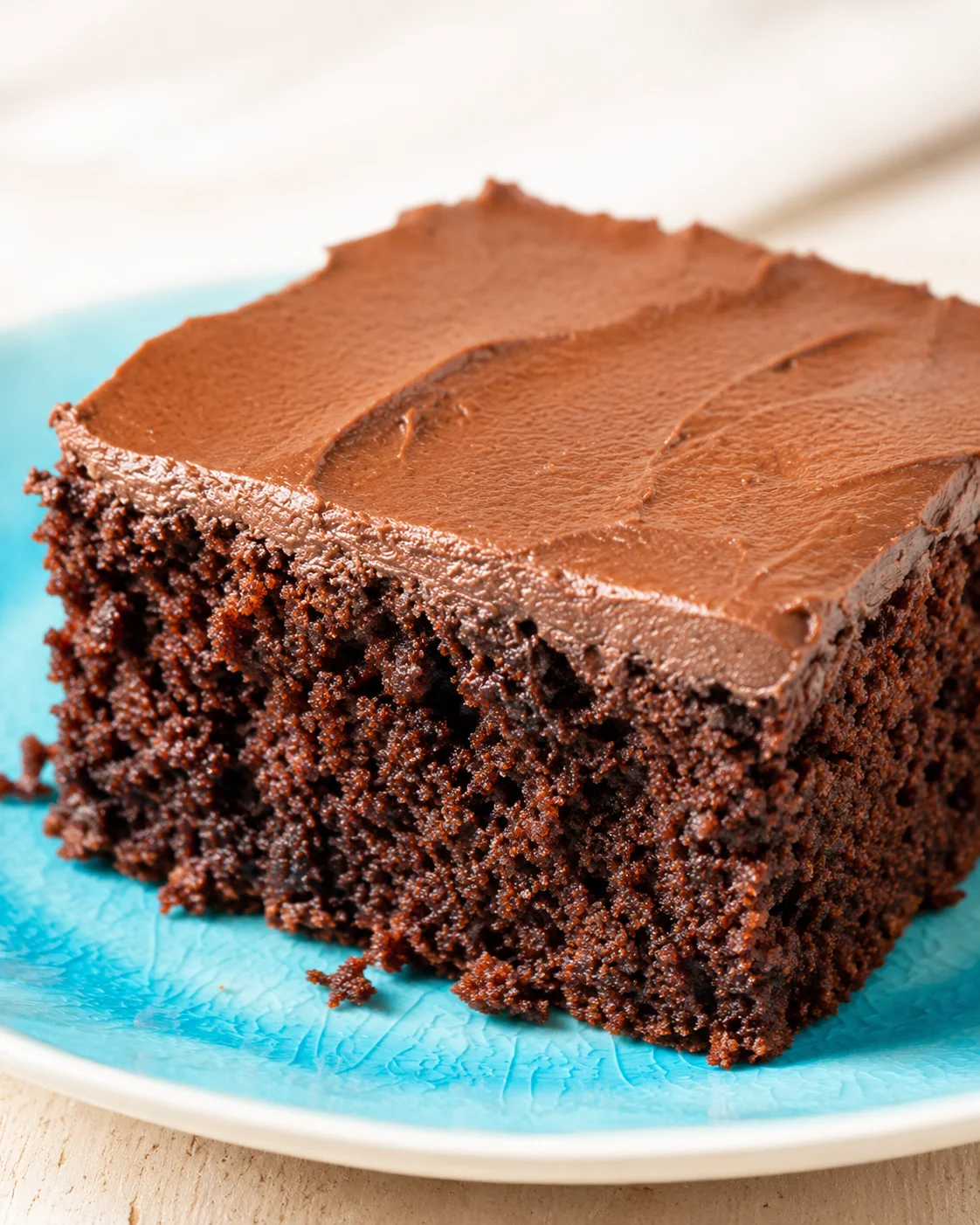Close-up of fudgy chocolate brownie on a blue plate, shows moist crumb and glossy top.
