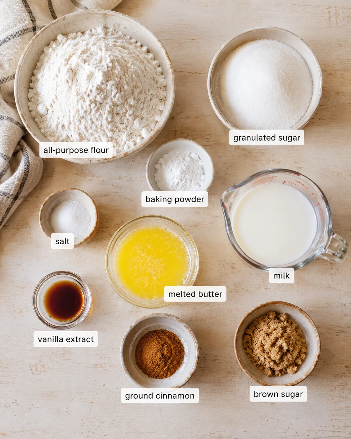 Top-down view of labeled baking ingredients on a light wooden surface, including flour, sugar, milk, butter, vanilla, salt, cinnamon, and brown sugar.