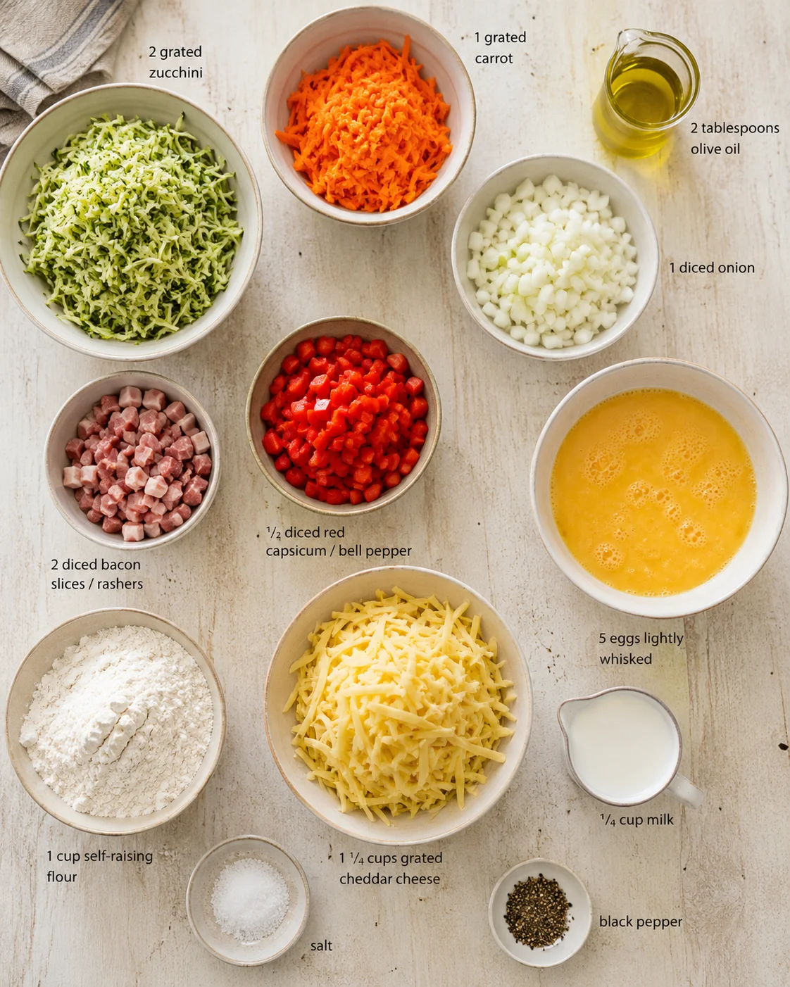 Overhead view of bowls with zucchini, carrot, onion, pepper, bacon, cheese, eggs, flour, milk, oil.
