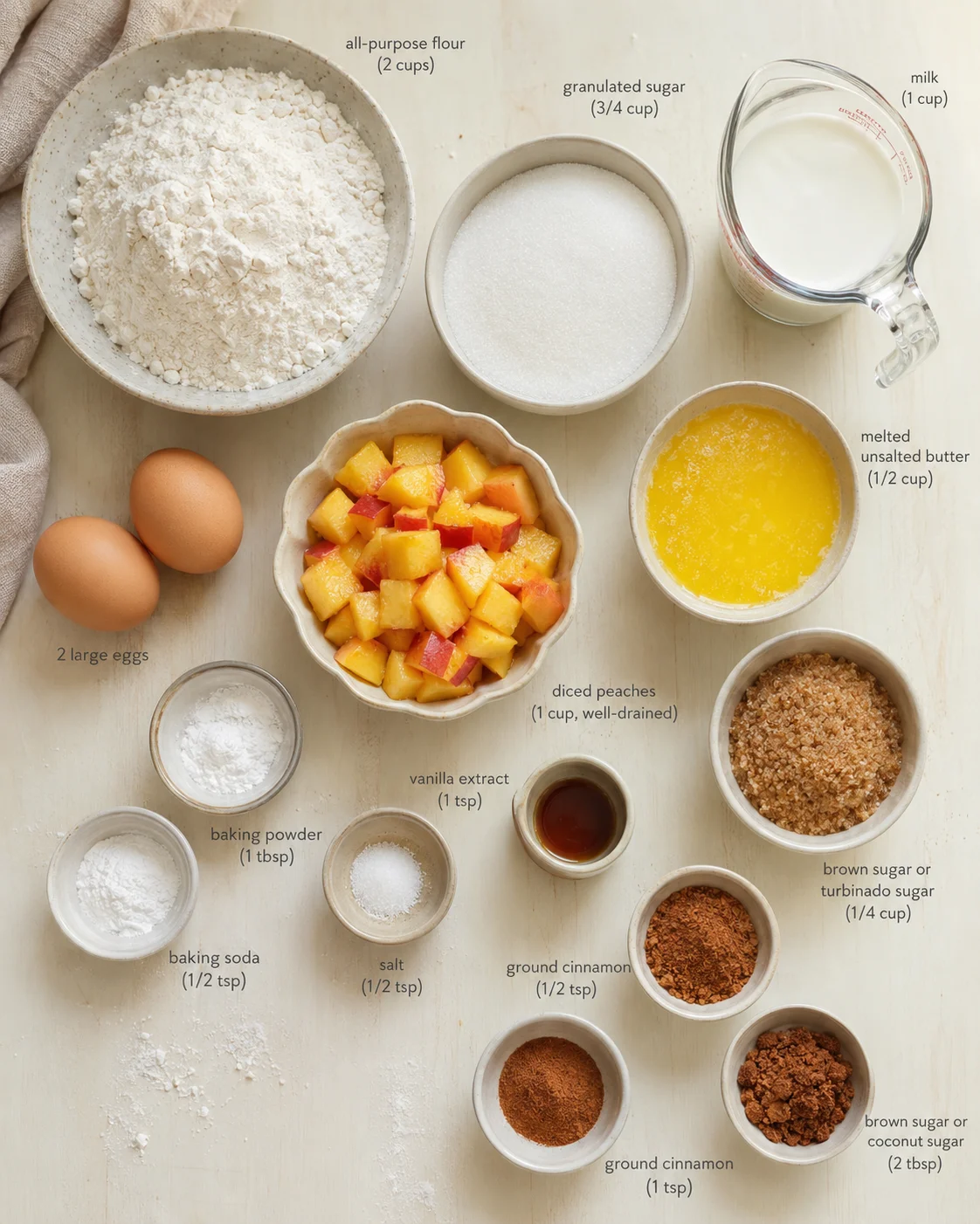 Flat lay of peach cake ingredients on a pale wooden surface, including flour, peaches, eggs, sugar, and butter