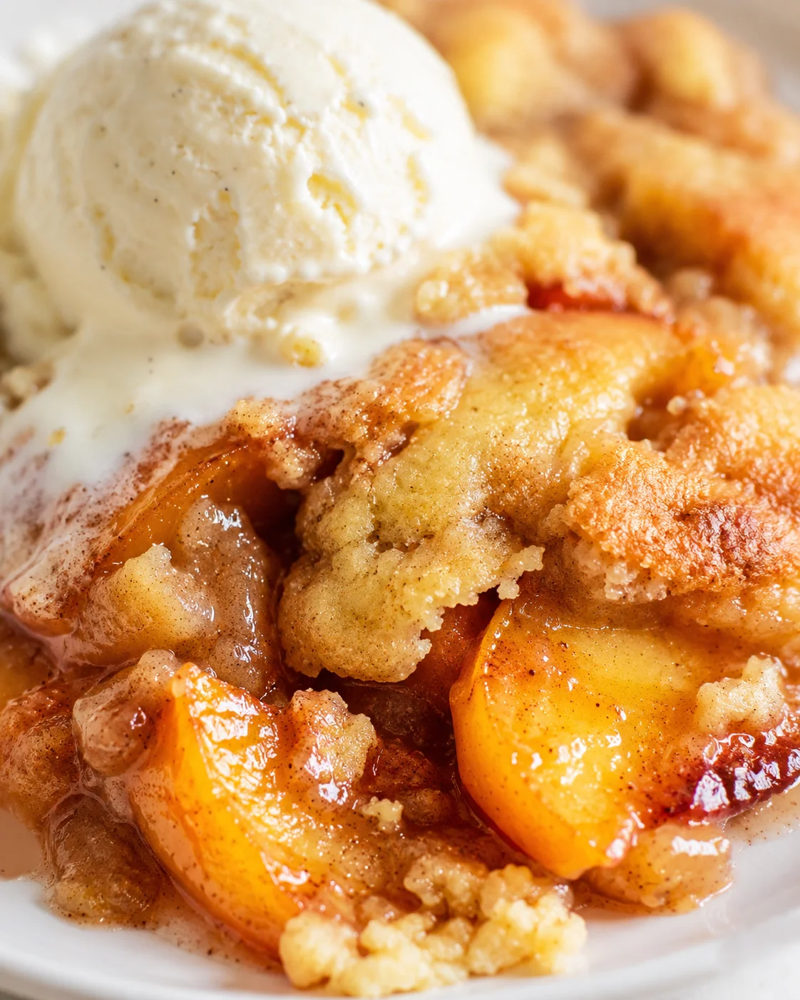 Close-up of peach cobbler with vanilla ice cream on a white plate, caramelized peaches visible.