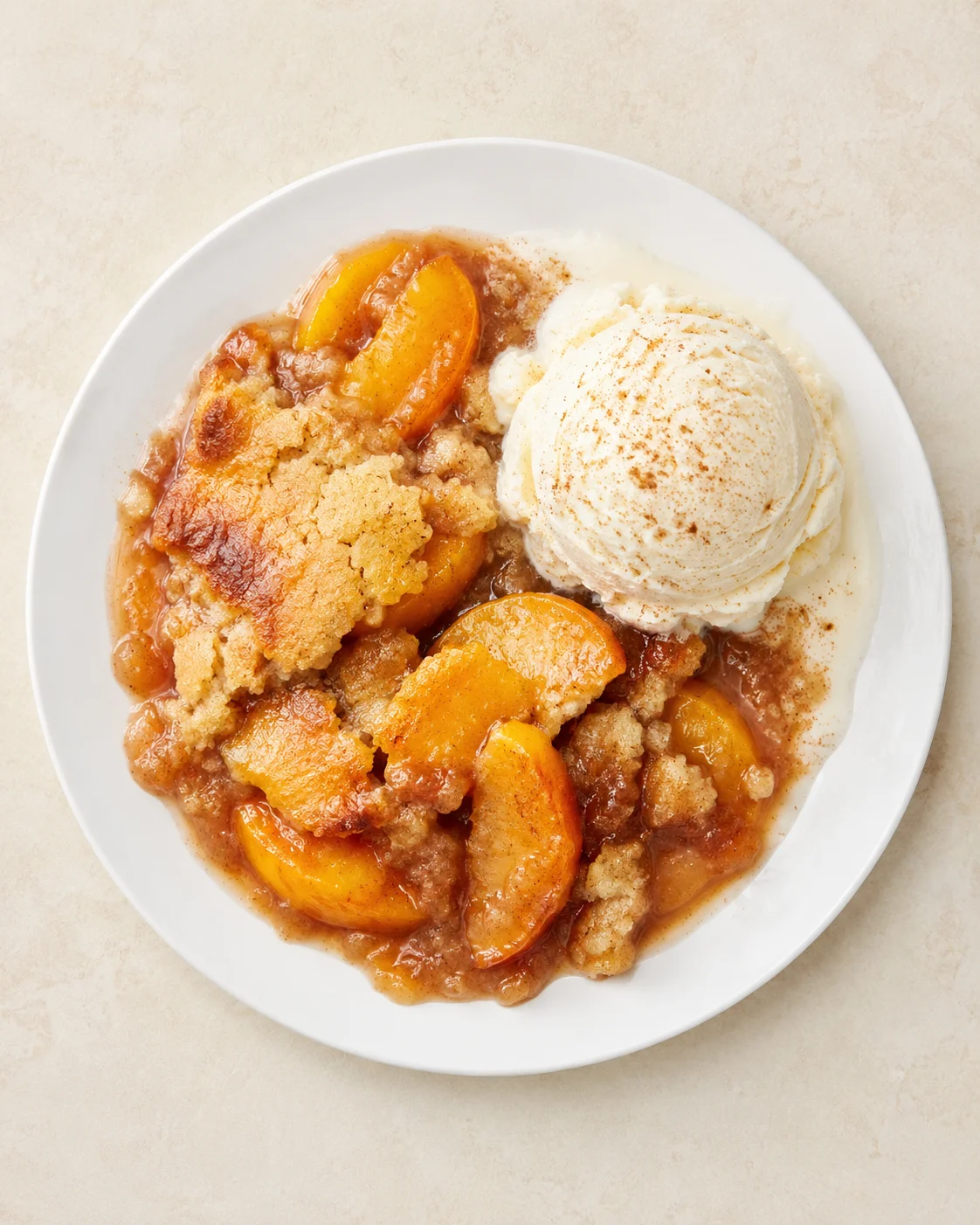 A round white plate holds peach cobbler with a scoop of vanilla ice cream, cinnamon dusting.