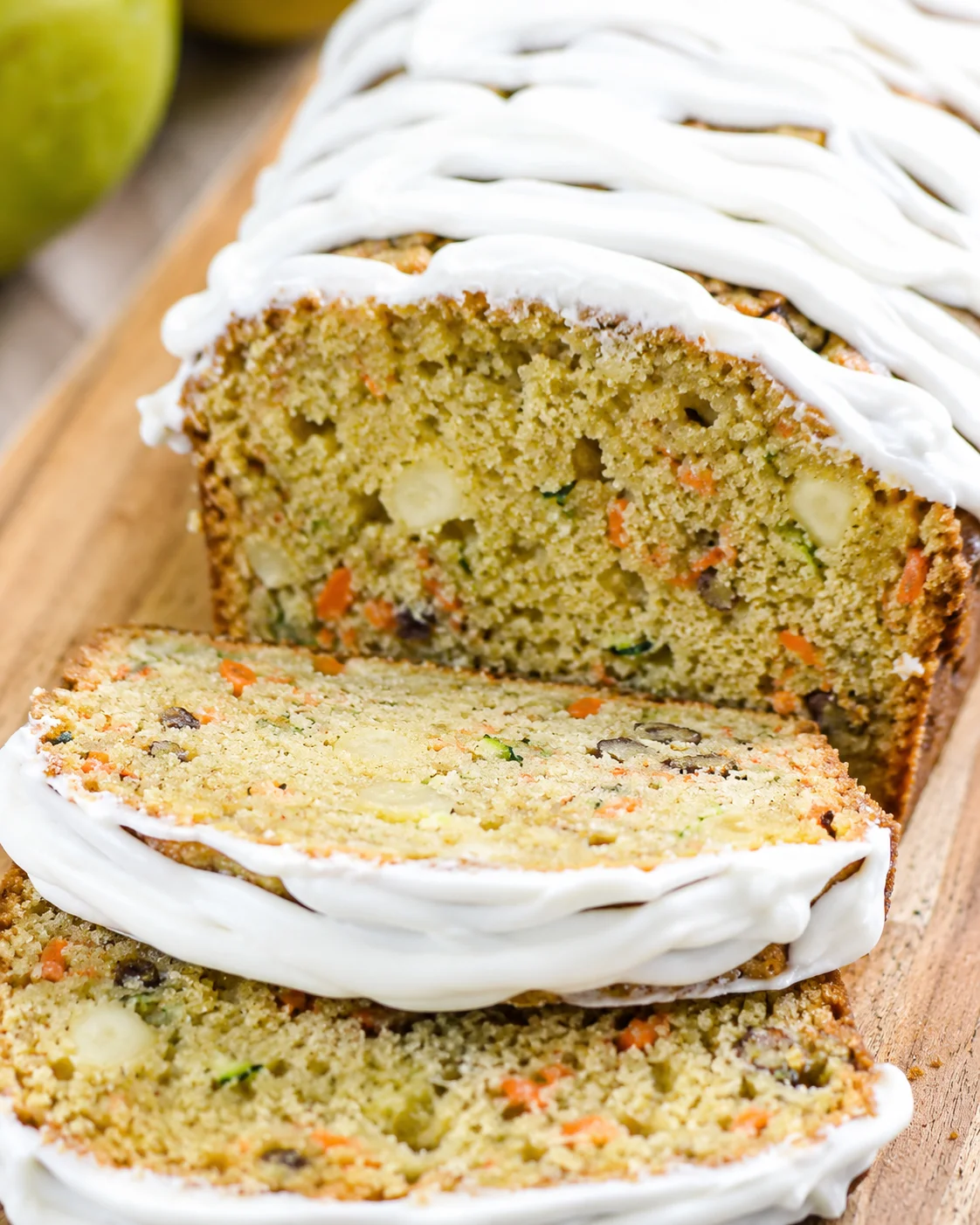 Slice of carrot-zucchini loaf cake with white cream-cheese frosting on a wooden board.