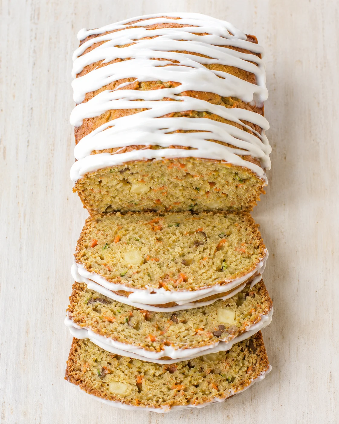 Loaf cake with white glaze, sliced to show carrot-vegetable crumb on a light wooden backdrop.