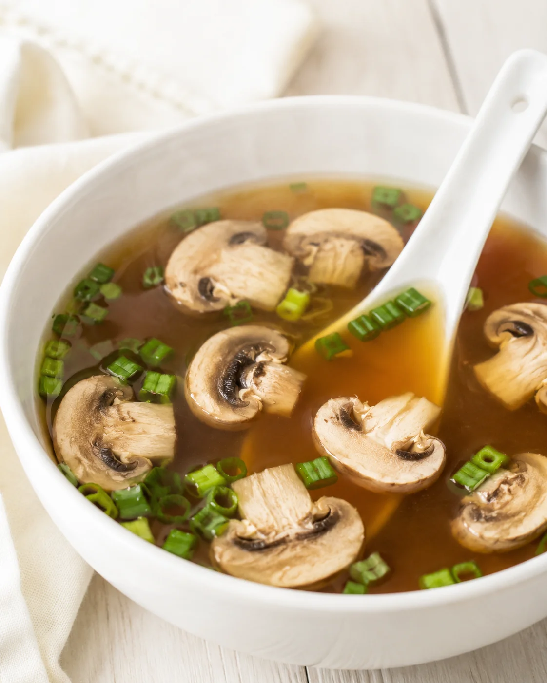 White bowl of mushroom soup with sliced mushrooms and green onions on a light wooden surface.