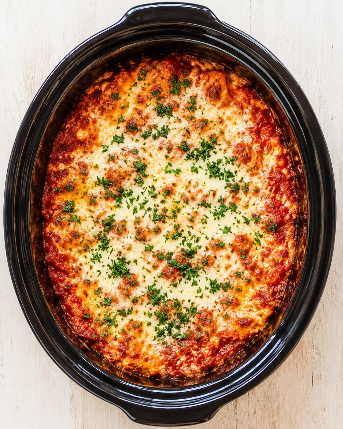 top-down view of cheese-topped lasagna in a black slow cooker, garnished with chopped parsley