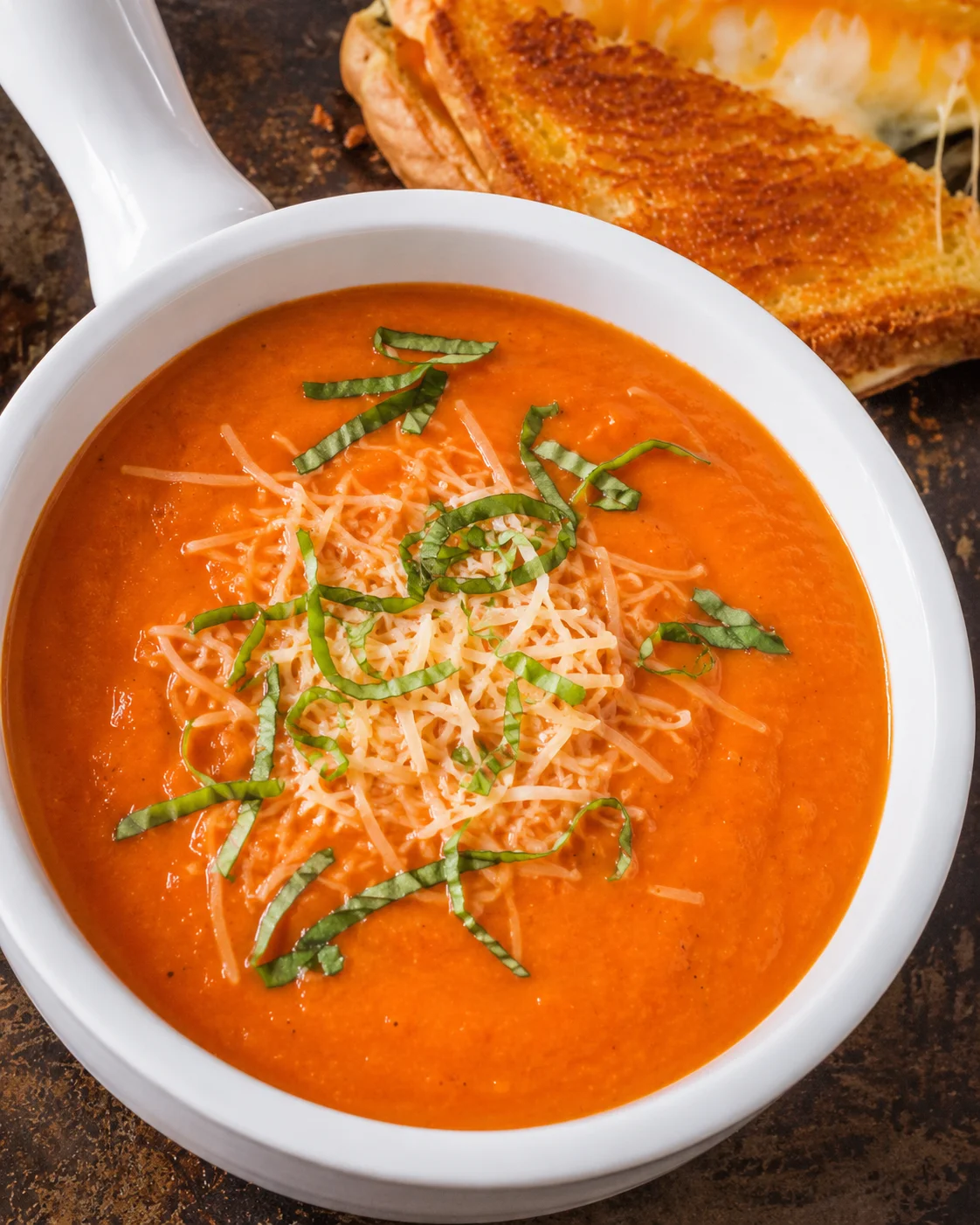 Bowl of creamy tomato soup topped with shredded parmesan and basil with a grilled cheese in the background
